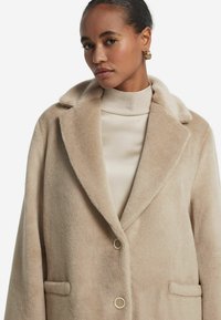 Beige Wollmischungsmantel mit Reverskragen, Drei-Knopf-Verschluss und Seitentaschen, mit einer weichen, strukturierten Oberfläche.