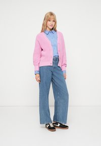 Cardigan rosa tricotado, camisa de botões azul às riscas, jeans de perna larga em denim, ténis preto e branco, de pé sobre um fundo branco.
