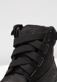 Black high-top boot made of suede and textile, featuring thick black laces, padded collar, and branding detail on the tongue.