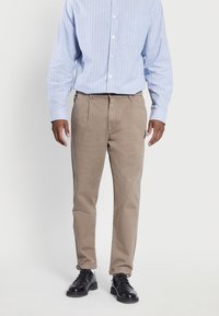 Man in beige plooibroek, zwarte leren schoenen, en een blauw-wit gestreept overhemd met lange mouwen, staand tegen een witte achtergrond.