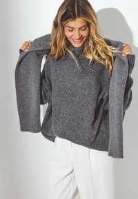 Suéter gris de punto con un corte relajado, puños acanalados y un cuello alto, usado sobre pantalones blancos. Textura suave y diseño casual.