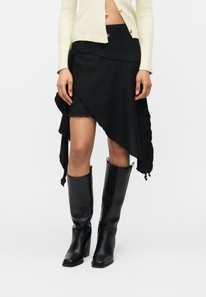 Nő fekete, aszimmetrikus fodros szegélyű szoknyában, térd fölé érő fekete bőrcsizmával és krémszínű, rövidített kardigánnal.