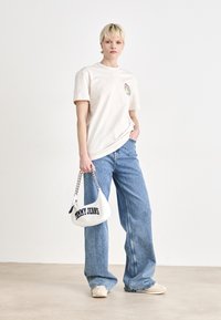 Šviesiai rudos medvilnės marškinėliai su spalvingu logotipu ant krūtinės, derinami su šviesiai mėlynomis plačiomis kelnėmis ir baltu rankiniu krepšiu su tamsiai mėlynu užrašu.