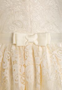 Koronkowa sukienka w kolorze kości słoniowej z misternymi kwiatowymi wzorami, fakturowaną górą i satynowym paskiem z kokardą w talii. Rozkloszowana spódnica poniżej.