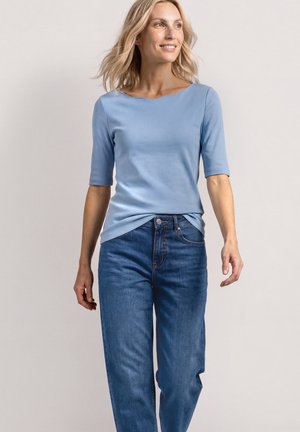 Femme souriante, marchant, portant une chemise ajustée bleu clair avec des manches aux coudes et un jean bleu coupe décontractée.