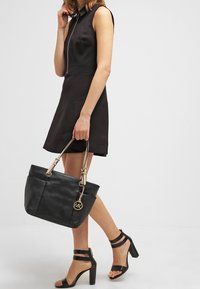 Svart läder tote-väska med guldfärgad hårdvara, beige remmar och en logocharm. Slät textur, rymligt inre och en strukturerad form.