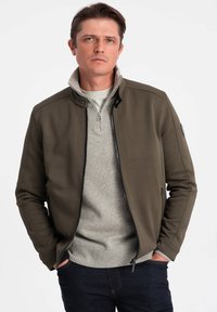 Giacca bomber verde oliva con superficie testurizzata, chiusura con zip e tasche laterali, abbinata a un maglione grigio chiaro sottostante.