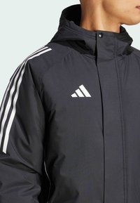 Schwarze Jacke mit weißem Dreistrich-Design auf den Schultern und Adidas-Logo. Verfügt über eine Kapuze und einen Frontsnappverschluss. Glattes, leichtes Material.