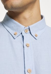 Camicia blu chiaro con colletto a punta, caratterizzata da bottoni in legno e un tessuto strutturato. La ripresa ravvicinata mostra i dettagli del collo e dei bottoni.