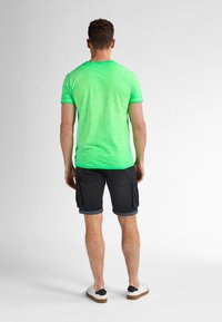 Tricou scurt, verde, purtat cu pantaloni scurți cargo negri. Tricoul are o finisare texturată; pantaloni scurți au manșete răsucite și buzunare laterale.