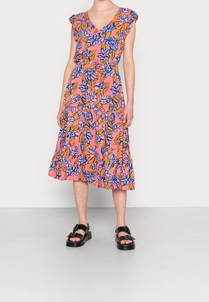 Kobieta w różowej sukience midi z niebiesko-żółtym kwiatowym wzorem, w czarnych sandałach na platformie, stojąca na jednolitym tle.