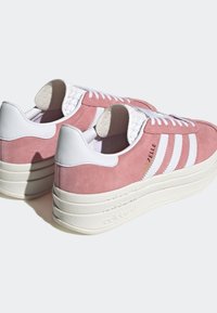Zapatillas Adidas Gazelle de ante rosa con rayas blancas, suelas de plataforma gruesas blancas y cierre de cordones blanco, mostradas desde los ángulos trasero y lateral.