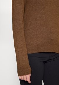 Brauner gestrickter Pullover mit gerippten Bündchen, mit einer strukturierten Oberfläche und einem lockeren Sitz. Kombiniert mit schwarzen Hosen, die die Länge des Kleidungsstücks zeigen.