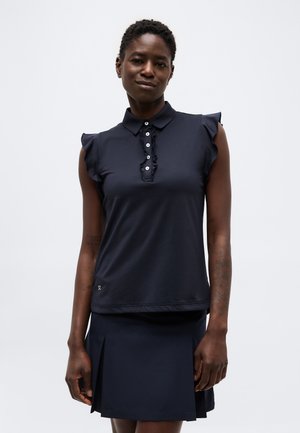 Naine, kes kannab mereväe sinist varrukateta polot särki volditud nööbitihendiga ja sobivat plisseeritud seelikut, seisab ühevärvilise taustaga.