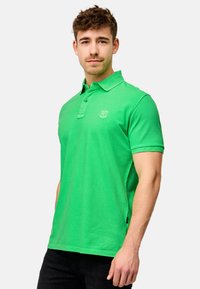 Grønn poloshirt laget av bomull, med krage, to-knapps placket og en liten logo på venstre bryst. Enkelt design med korte ermer.