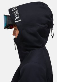 Giacca esterna nera con colletto alto, cappuccio regolabile con cordino e logo "Peak Performance". Presenta un design elegante e un tessuto testurizzato.