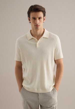 Jonge man die een crèmekleurig geribd poloshirt met korte mouwen en gestreepte broek draagt, staand met zijn handen in de zakken tegen een effen achtergrond.