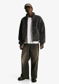 Dark gray zip-up fleece jacket with textured pattern, white t-shirt, light brown wide-leg jeans, and black shoes. Cap with checkered design.