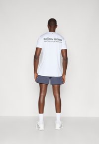 Λευκό αθλητικό μπλουζάκι με την ένδειξη "BJÖRN BORG TRAINING CLUB MONACO" σε σκούρο πράσινο χρώμα, συνδυασμένο με ναυτικά σορτς και λευκά αθλητικά παπούτσια σε ουδέτερο φόντο.