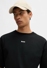 Czarny t-shirt z okrągłym dekoltem z białym logo "BOSS" na klatce piersiowej; materiał bawełniany; zestawiony z jasnobeżową czapką z kwadratową naszywką logo.