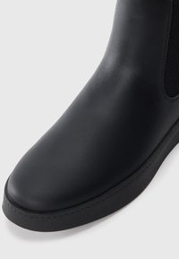 Botín negro con material de cuero suave, punta redondeada y panel lateral elástico. Suela de goma texturizada con un sutil detalle de costura.