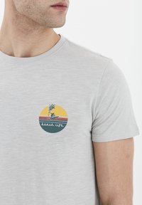 Lys grå bomull T-skjorte med en sirkulær grafikk av en solnedgang, palme og teksten "strandliv" i kontrastfarger.