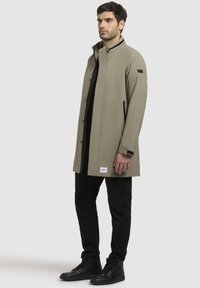 Khaki lang frakke med høj krave, trykknap lukning foran, sidelommer med lynlås; parret med sorte bukser og sneakers; glat stof.
