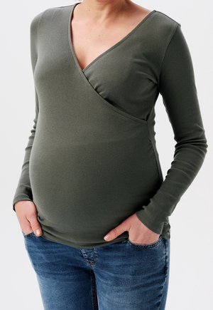 Osoba w ciąży ubrana w oliwkowo-zieloną bluzkę z długim rękawem i niebieskie dżinsy, z rękami spoczywającymi w przednich kieszeniach, na jednolitym tle.