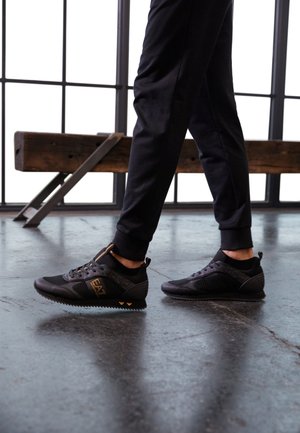 Personne portant des baskets noires avec logo doré EA7 et un pantalon de jogging noir marchant sur un sol en béton poli près d'un banc en bois.