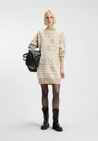 Beige neulemekko, jossa on valkoinen "HUGO"-logokuosi, korkea kaulus ja pitkät hihnat, yhdistettynä mustaan teksturoituun laukkuun ja polven yli ulottuviin saappaisiin.
