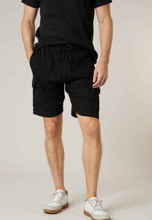 Man draagt zwarte casual korte broek met zijzakken, een zwart shirt met korte mouwen en witte sneakers met lichtbruine zolen.