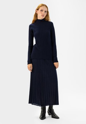 Marineblauer, gerippter Strickpullover mit hohem Kragen, kombiniert mit einem plissierten, langen Rock. Strukturierten Stoff, tailliertes Oberteil und ausgestellter Saum. Schwarze Knöchelstiefel.