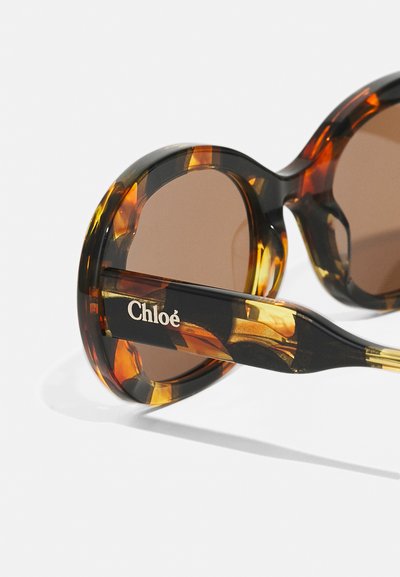 Okrągłe okulary przeciwsłoneczne w oprawie w panterkę, z brązowymi soczewkami i logo "Chloé" na zauszniku.