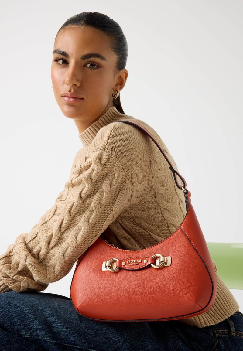 Czerwona skórzana torba hobo z złotymi detalami i kontrastującymi akcentami, z logo i wygiętą sylwetką. Teksturowany sweter z dzianiny w kolorze beżowym.