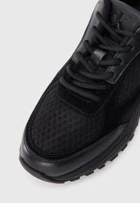 Scarpa sportiva nera con tomaia in rete e dettagli in suede. Punta rotonda, lacci neri piatti e suola in gomma texturizzata per una presa ottimale.