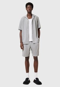 Lys grå kortærmet skjorte med knapper, matchende shorts, minimalistisk design, parret med sorte slip-on sko og hvide sokker. Blød tekstur.
