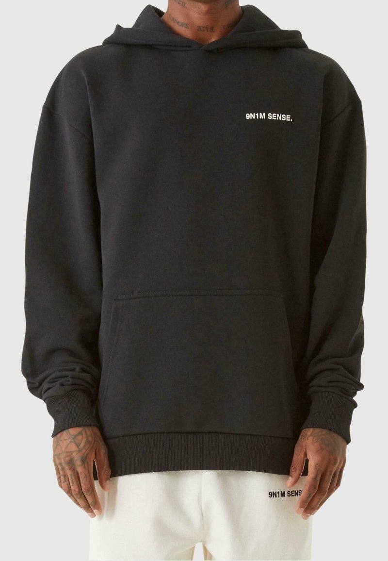 Homme portant un sweat à capuche noir avec le texte "9N1M SENSE." sur la poitrine et un pantalon blanc avec le même texte sur la cuisse gauche.