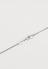 Collier en chaîne argentée avec une étiquette rectangulaire gravée "ICON" et un fermoir à ressort, sur un fond blanc uni.
