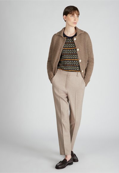 Brązowy sweter z dużymi guzikami noszony na wzorzystym czarnym, żółtym i niebieskim swetrze, w połączeniu z jasnobeżowymi, dopasowanymi spodniami i ciemnymi butami.