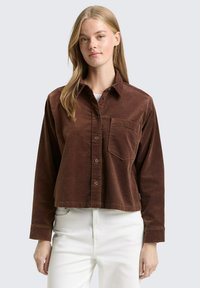 Bruin corduroy cropped shirt met een puntige kraag, knopenluik aan de voorkant, één borstzak en lange mouwen, gedragen over witte broek.
