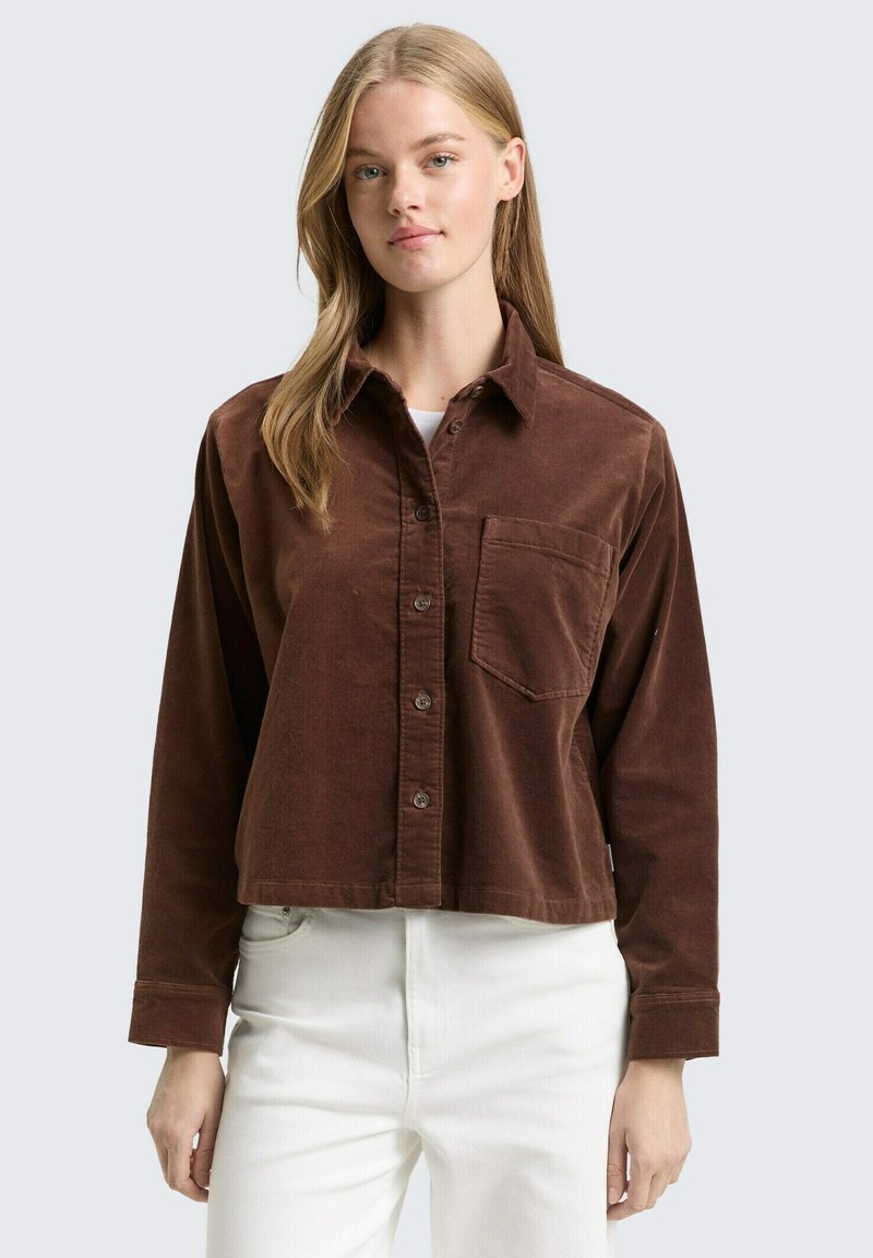 Bruin corduroy cropped shirt met een puntige kraag, knopenluik aan de voorkant, één borstzak en lange mouwen, gedragen over witte broek.