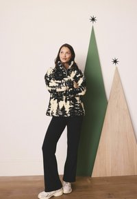 Pulover din fleece de culoare crem cu un model de siluetă de copac negru, guler înalt, asortat cu pantaloni negri evazați și pantofi sport crem.