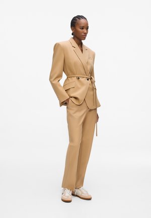 Vrouw met gevlochten haar die een beige blazer met riem draagt, bijpassende broek en beige witte sneakers, staand met handen in de zakken.