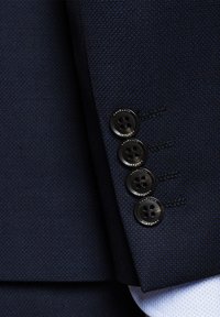 Rękaw granatowego żakietu z teksturowanej tkaniny, czterema czarnymi guzikami i subtelnymi detalami szycia w pobliżu mankietu.