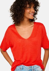 Red short-sleeve v-neck top in lightweight fabric, featuring a relaxed fit and subtle texture. Paired with light blue denim shorts.