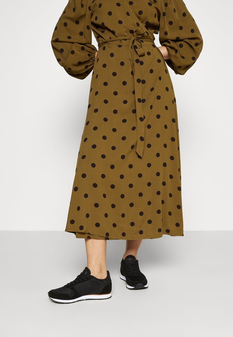Femme portant une robe midi vert olive à pois noirs avec des manches bouffantes, associée à des baskets noires, debout sur un sol gris clair.