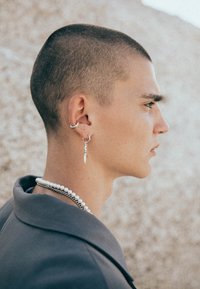 Silverörhänge, inklusive en hoop och en dolkdesign, som bärs av en person med kort hår. Lager av pärl- och kedjearmband, grå blazer.