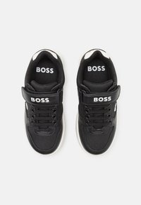 Schwarz-weiße Kindersneaker mit Schnürsenkeln und Klettverschluss, mit dem Markennamen "BOSS" auf dem Klettverschluss und der Innensohle, von oben auf einem weißen Hintergrund betrachtet.