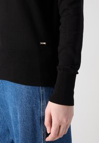 Svart stickad tröja med ribbad mudd och fåll, pryds av en liten silverdetalj. Matchad med ljusblå denimbyxor.