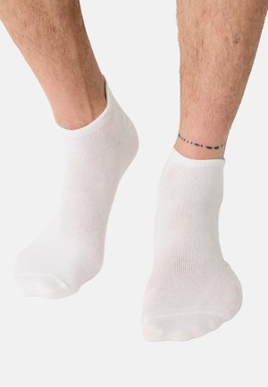 Beine mit schlichten weißen Knöchelsocken, ein Fuß leicht angehoben, mit einem gepunkteten Linien-Tattoo um den rechten Knöchel vor hellem Hintergrund.